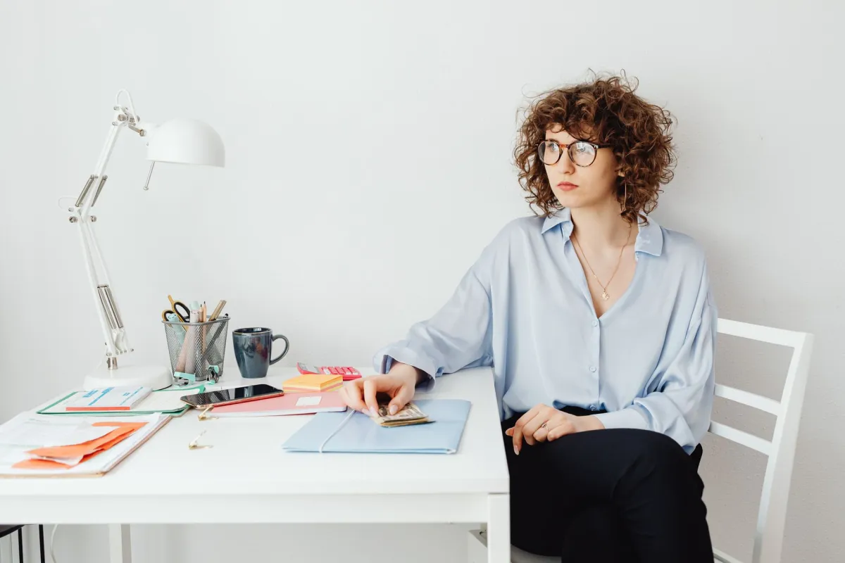 blouse, blue, bookkeeper, bookkeeping, bookkeeping_accounting, business, commercial, company, corporate, counting cash, curly hair, desk, desk organizer, enterprise, financial, folder, glasses, office, office supplies, planner, professional, receipts, sitting, stationery, woman, workspace
